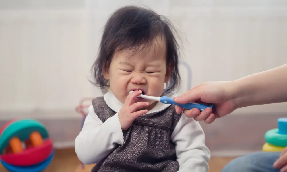 asian toddler making an angry face as parent tries to brush her teeth hwo to make brushing infant teeth easier